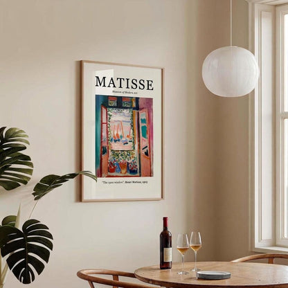 A framed art print of Henri Lebasque's Matisse 'The Open Window' poster hanging on a beige wall in a modern dining room setting with a wooden table, wine bottle, and glasses, alongside a white globe pendant light and a green monstera plant.