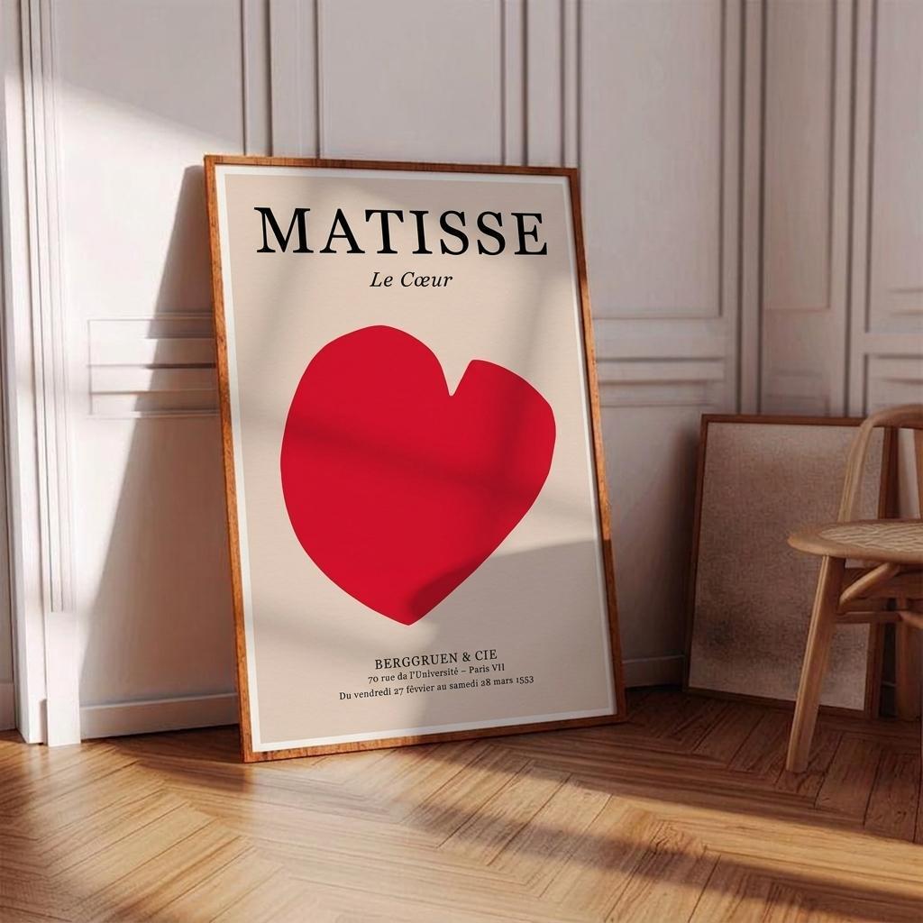 A framed art print leaning against a white paneled wall on a wooden floor. The print features the word 'MATISSE' in black serif letters above a large red heart on an off-white background, with smaller black text below.