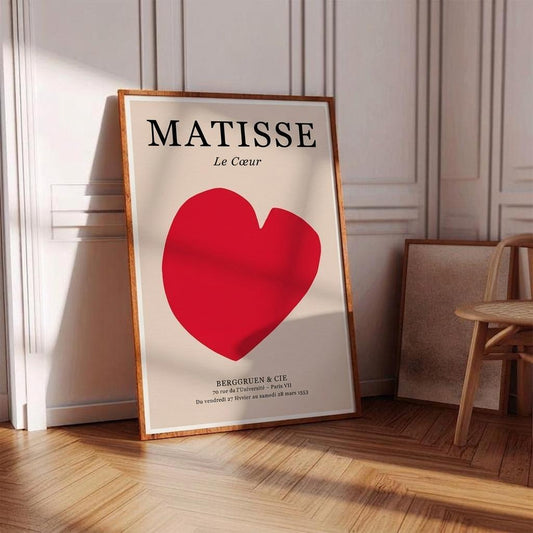 A framed art print leaning against a white paneled wall on a wooden floor. The print features the word 'MATISSE' in black serif letters above a large red heart on an off-white background, with smaller black text below.