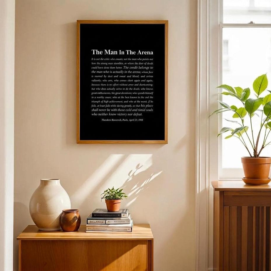 A framed black art print with white text reading 'The Man In The Arena' hangs on a beige wall in a styled interior. Below the print is a wooden sideboard with a cream vase, small books, and a potted plant, while a larger plant sits by a window on the right.