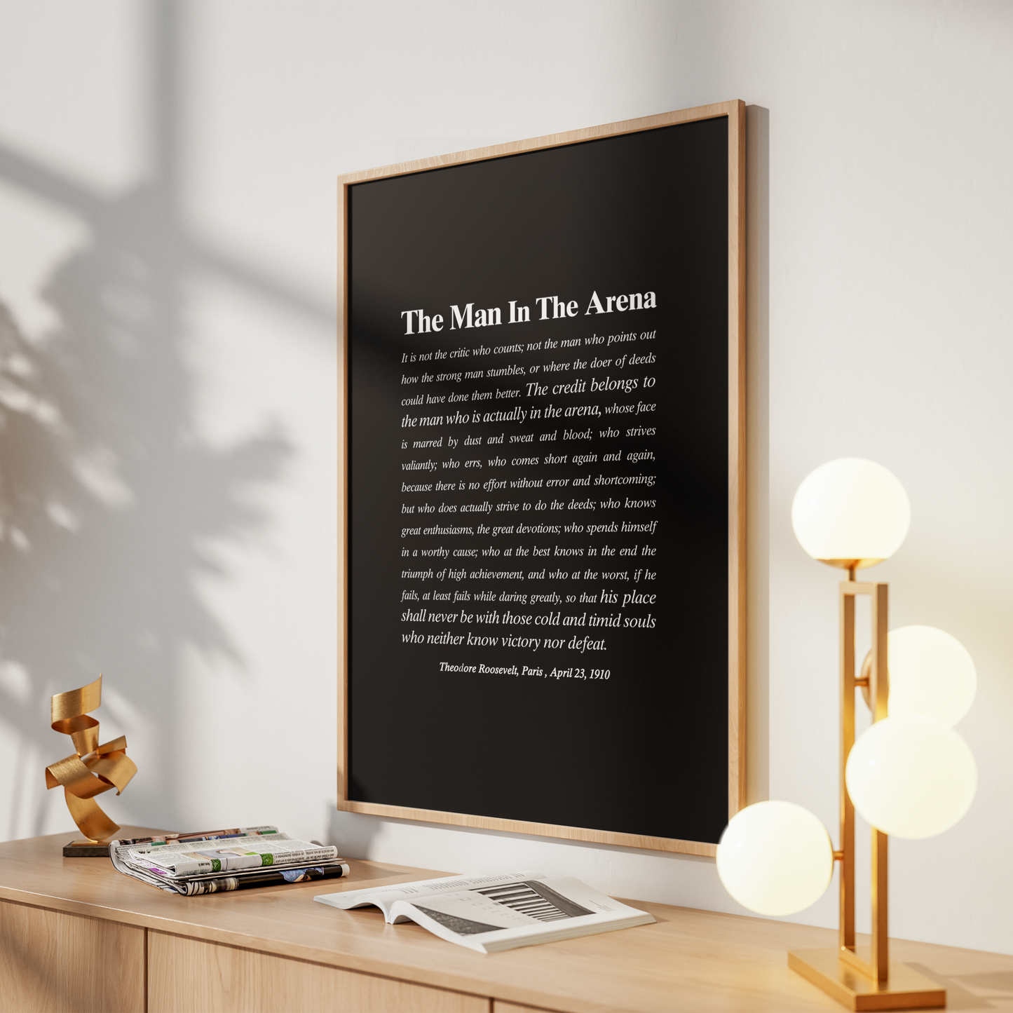 A framed vertical art print featuring a quote from 'The Man In The Arena' by Theron Ramsay, displayed on a wooden console table in a modern living room setting with a lamp, books, and a sculpture.