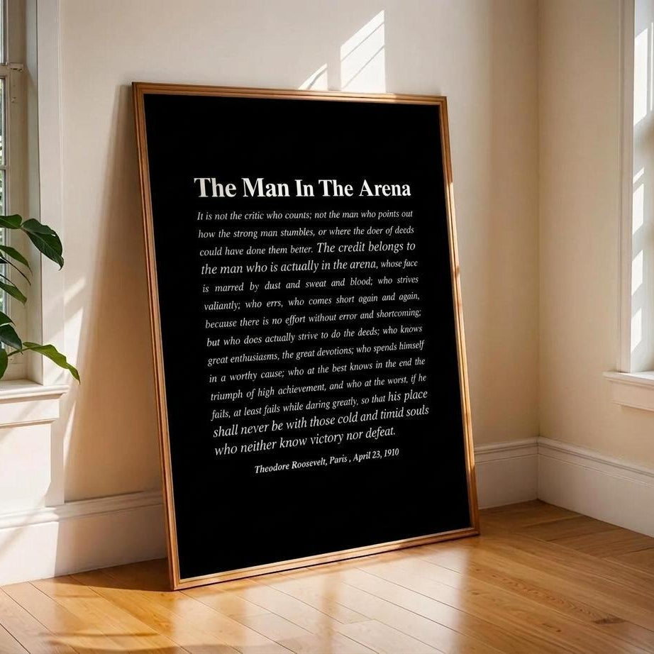 A framed black art print titled 'The Man In The Arena' with white text, leaning against a white wall on a wooden floor in a sunlit room with windows and a plant.