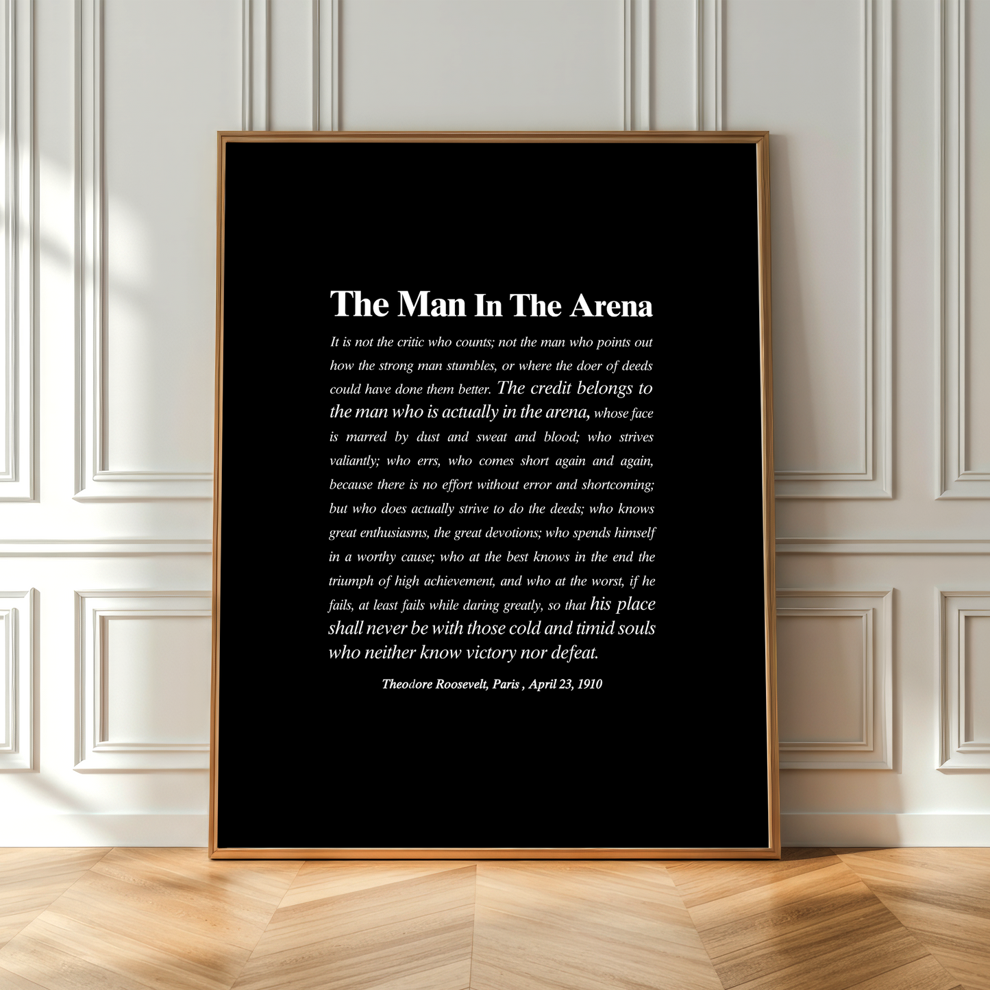 A framed black art print featuring a white quote titled 'The Man In The Arena' by Theodore Roosevelt, dated April 23, 1903, leaning against a white paneled wall on a light wood floor.