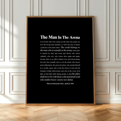 A framed black art print featuring a white quote titled 'The Man In The Arena' by Theodore Roosevelt, dated April 23, 1903, leaning against a white paneled wall on a light wood floor.