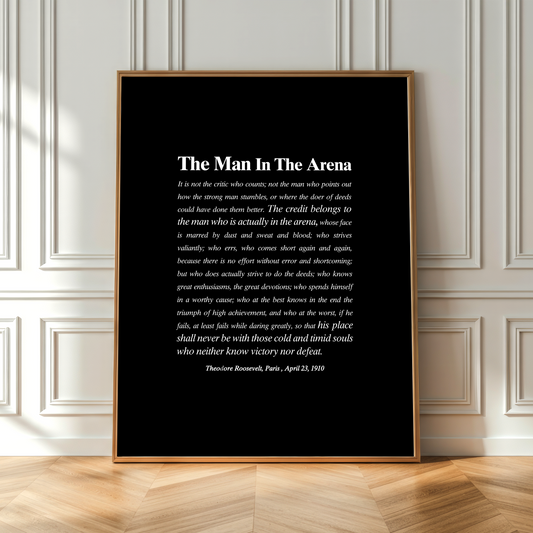 A framed black art print featuring a white quote titled 'The Man In The Arena' by Theodore Roosevelt, dated April 23, 1903, leaning against a white paneled wall on a light wood floor.