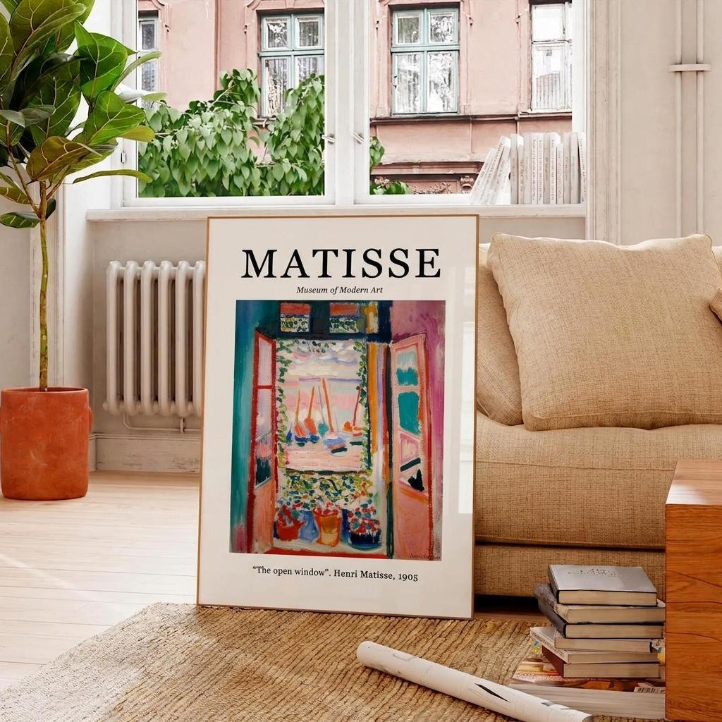 A framed art print of Henri Matisse's 'The open window' leaning against a beige sofa in a bright, modern living room with a potted plant and window in the background.