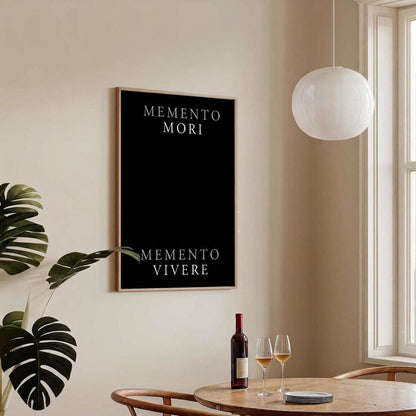 A framed wall art print with the Latin phrases 'MEMENTO MORI' and 'MEMENTO VIVERE' in white text on a black background, hanging in a warm beige dining room above a wooden table with wine glasses and a bottle.