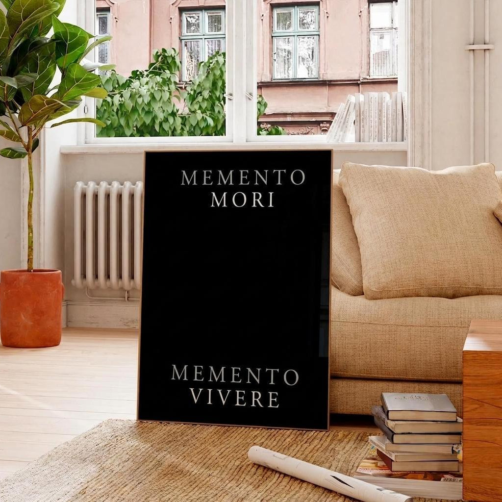 A black framed art print with the Latin phrases 'MEMENTO MORI' and 'MEMENTO VIVERE' in white serif lettering, displayed in a modern living room setting next to a beige sofa and a potted plant.