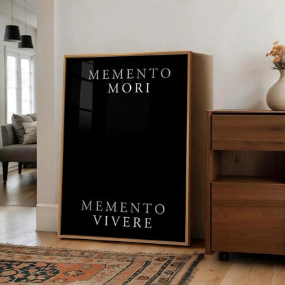 A large framed wall art print leaning against a white wall in a modern living room. The print has a black background with the Latin phrases 'MEMENTO MORI' at the top and 'MEMENTO VIVERE' at the bottom in white serif text, surrounded by a light wood frame.