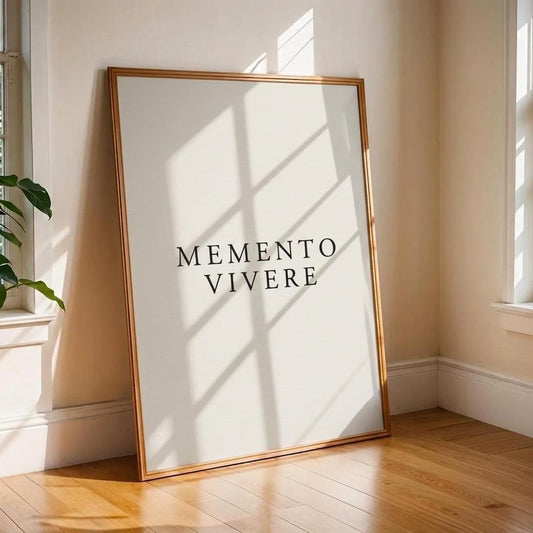 A framed minimalist art print with the text 'MEMENTO VIVERE' in black serif letters on a white background, leaning against a beige wall on a wooden floor in a sunlit room.