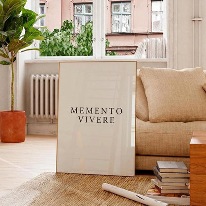 A vertical framed art print with the text 'MEMENTO VIVERE' in black serif font on a warm off-white background, leaning against a beige sofa in a bright, modern living room.