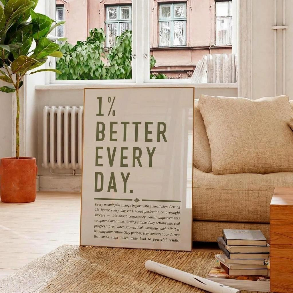 A vertical framed art print with the text '1% BETTER EVERY DAY.' in dark green on a cream background, styled in a cozy living room setting next to a beige sofa, a potted plant, and a stack of books.