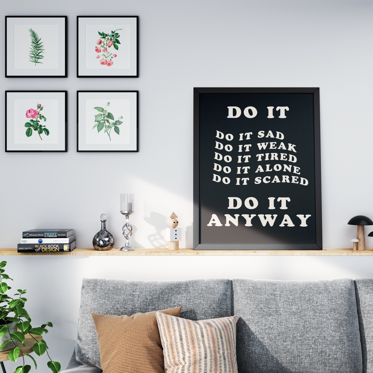 A framed typographic wall art print with the words 'DO IT ANYWAY' in white on a black background, displayed above a gray sofa in a styled living room. The room also includes four framed botanical prints on the wall, a wooden shelf with decor, and a plant in the foreground.