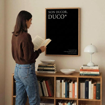 A woman stands in a room reading a book in front of a framed black poster with the text 'NON DUCOR, DUCO*'. The room includes a wooden bookshelf filled with books, white bowls, and a white table lamp against a warm beige wall.