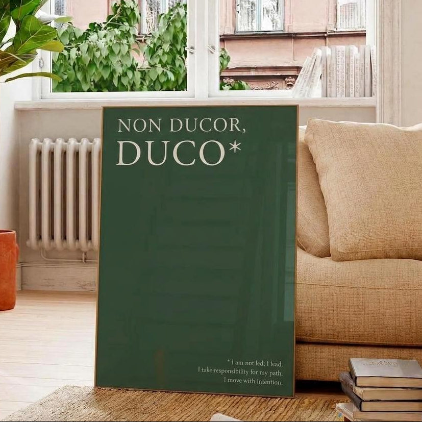 A large dark green book titled 'NON DUCOR, DUCO*' stands upright on a woven rug in a living room setting, with a beige sofa, potted plant, windows, and books visible.