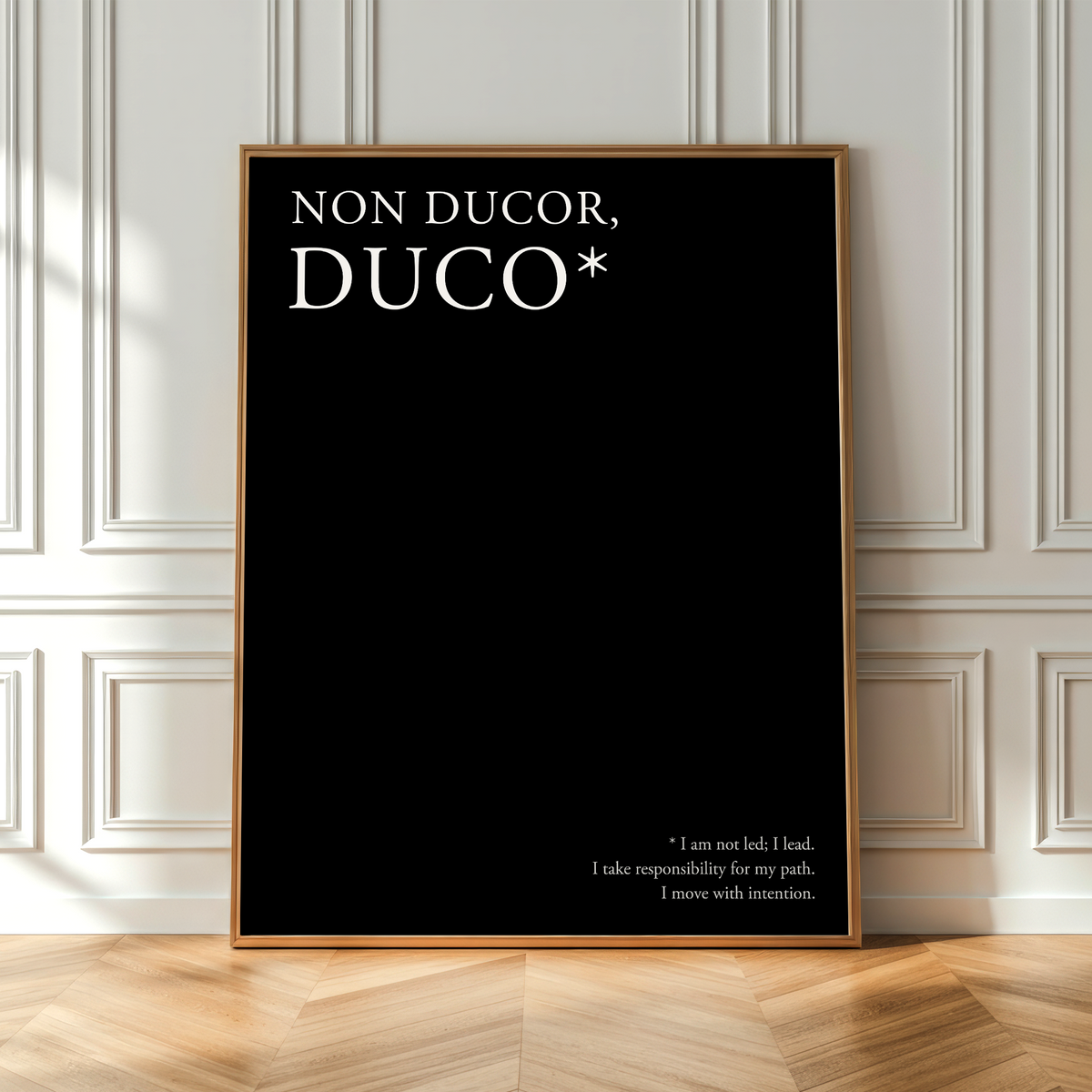 A framed black poster leaning against a white paneled wall on a light wood floor. The poster features the text 'NON DUCOR, DUCO*' in large white letters at the top and a smaller quote at the bottom right.