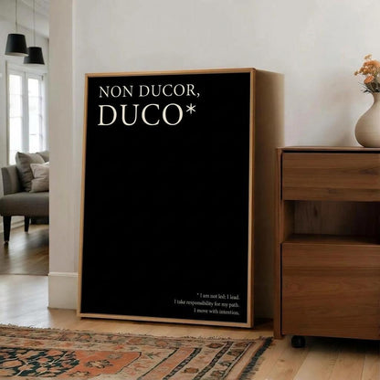 A framed black wall art print with the Latin phrase 'NON DUCOR, DUCO*' in white serif text, leaning against a white wall in a modern living room next to a wooden cabinet.