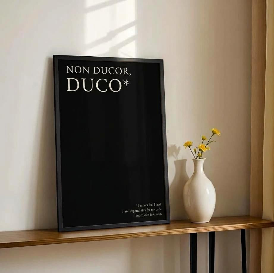 A black framed art print with the Latin phrase 'NON DUCOR, DUCO*' in white lettering, leaning on a wooden console table next to a white ceramic vase with yellow flowers against a beige wall with warm sunlight and curtains.