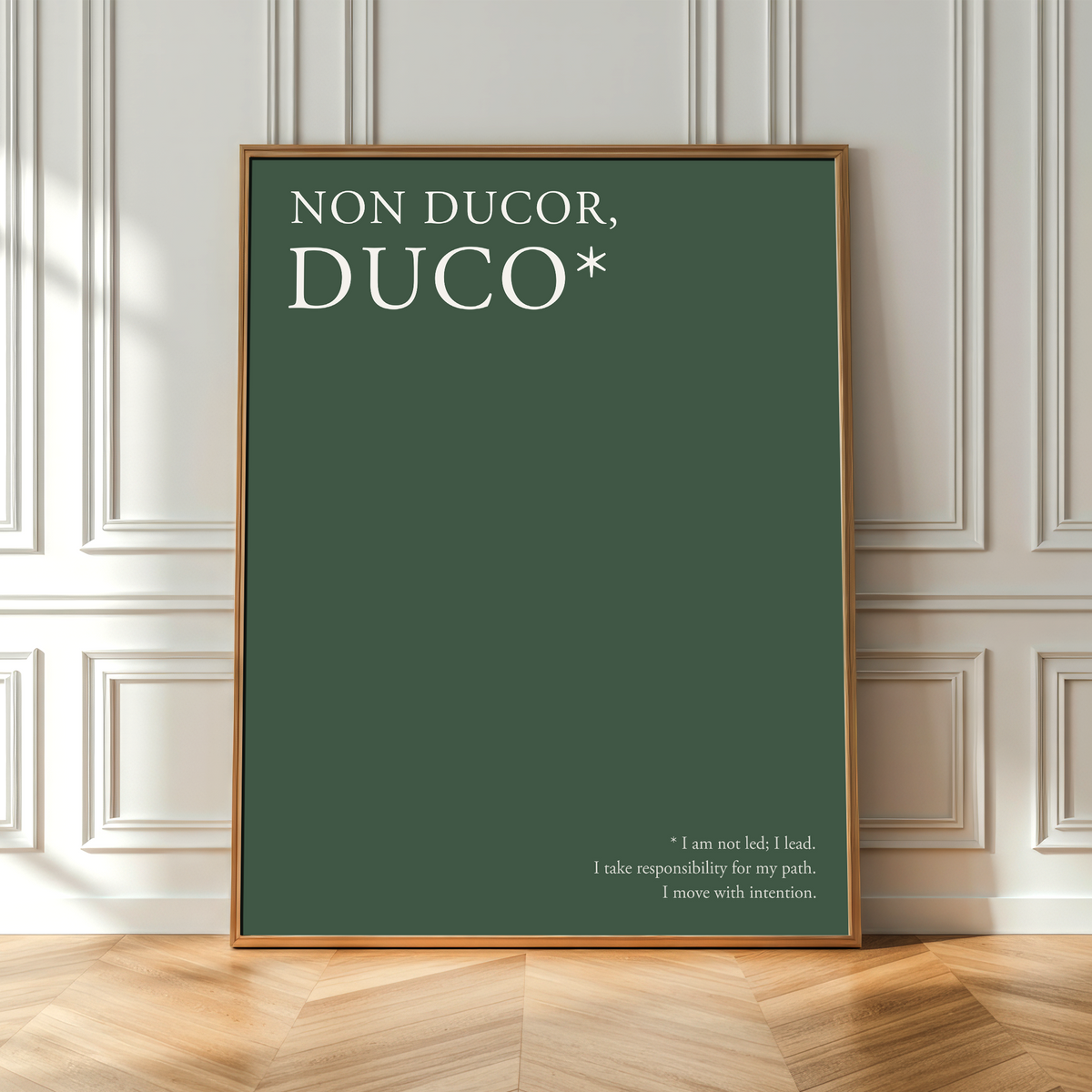 A framed art print with the Latin text 'NON DUCOR, DUCO*' in white on a dark green background, leaning against a white paneled wall on a light wood herringbone floor.