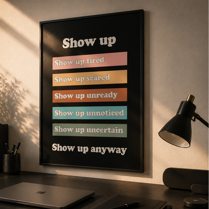 A framed motivational poster with the text 'Show up anyway' and colorful status bars hangs on a wall above a dark desk with a laptop, mug, lamp, and plant in a sunlit home office.