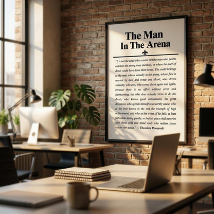 A framed vertical poster titled "The Man In The Arena" with a black text print on a white background, hanging on a brick wall in a modern office or cafe setting with blurred desks and chairs in the foreground.