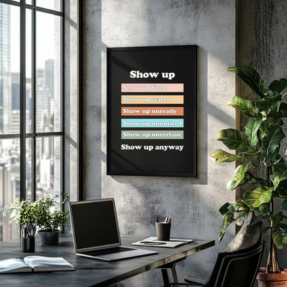 A framed wall poster with the text 'Show up' and stacked status lines in different colors hangs on a concrete wall above a desk with a laptop, plants, and a chair in a modern office setting.