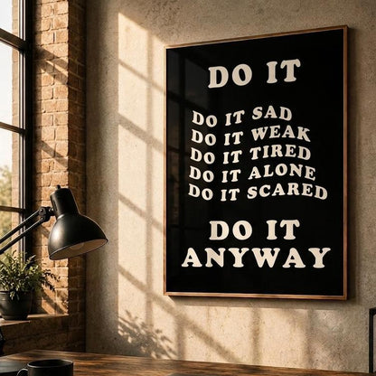 A framed motivational wall art print with the text 'DO IT ANYWAY' in white on a black background, hanging on a textured beige wall in a styled home office setting with a desk, lamp, and whiteboard.