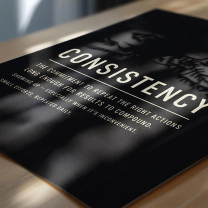 A black motivational poster with white text reading 'CONSISTENCY' and a quote about repeating right actions, photographed at an angle on a light wood surface.