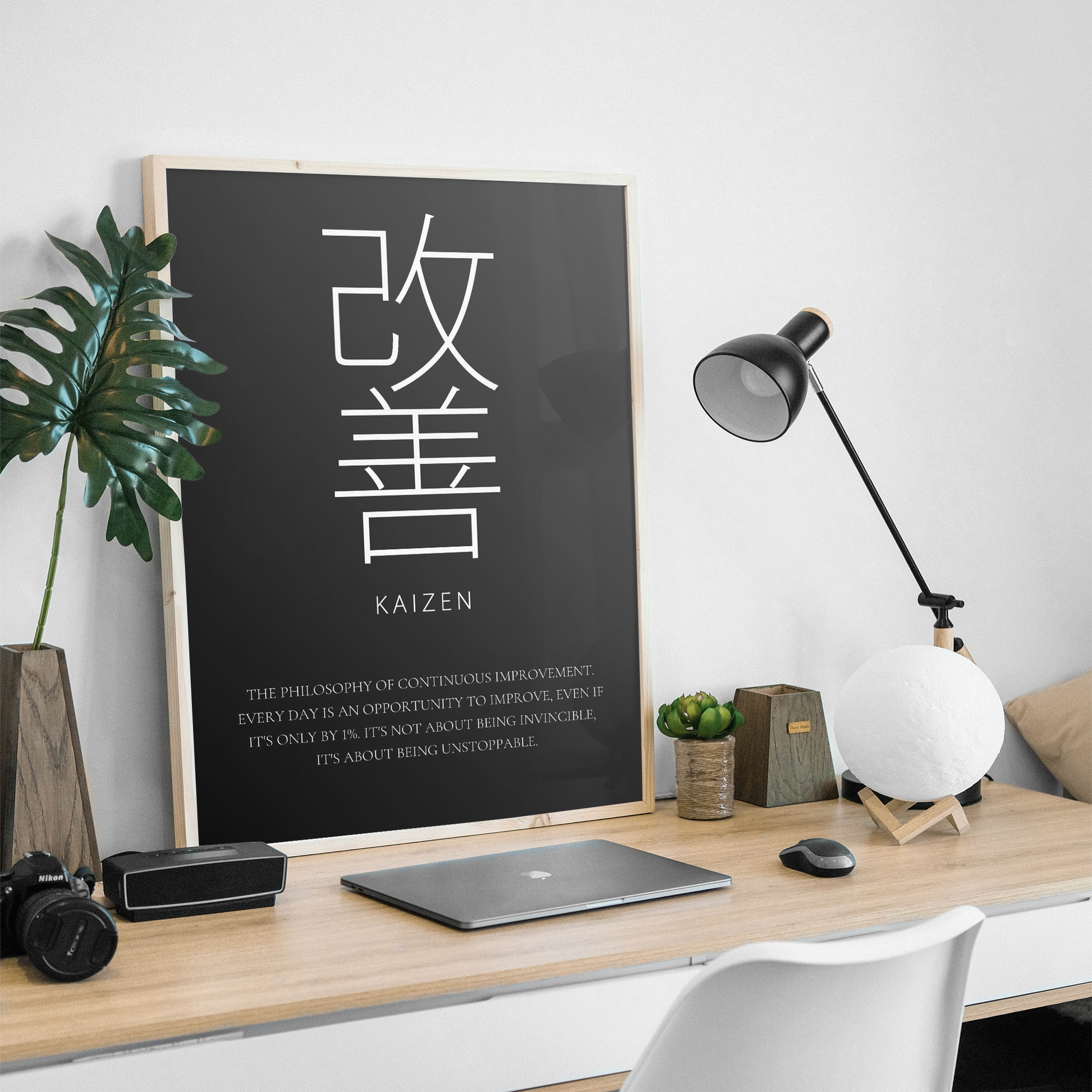 A framed black poster with large white Japanese characters and the English word KAIZEN hangs on a light wood desk in a styled home office setting. The scene includes a laptop, black desk lamp, white globe lamp, small plant, speaker, and a white chair against a white wall.