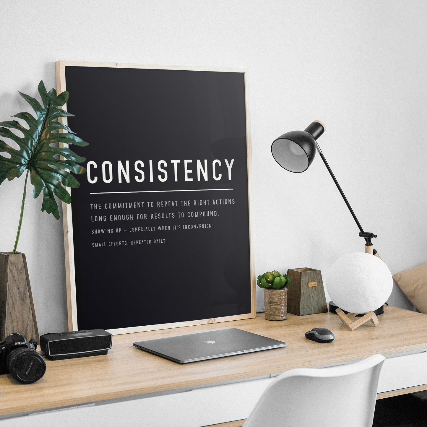 A framed black motivational poster with the word 'CONSISTENCY' in large white letters stands on a light wood desk in a styled home office setup. The scene includes a laptop, camera, speaker, plant, lamp, small succulent, and a white chair against a white wall.