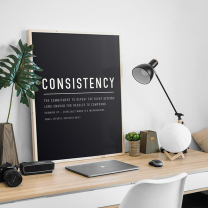 A framed black motivational poster with the word 'CONSISTENCY' in large white letters stands on a light wood desk in a styled home office setup. The scene includes a laptop, camera, speaker, plant, lamp, small succulent, and a white chair against a white wall.