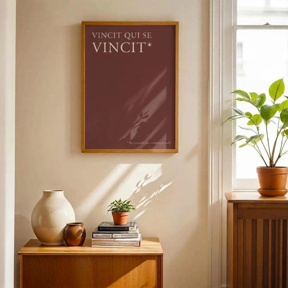 A framed art print featuring the French text 'VINCIT QUI SE VINCIT*' in white on a dark reddish-brown background, hanging on a beige wall in a sunlit room next to a window with a green plant.