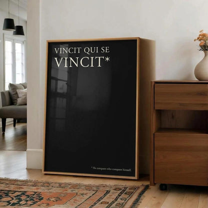 A framed black poster leaning against a white wall in a modern living room setting. The poster features the text 'VINCIT QUI SE VINCIT*' in white, with a small Latin phrase at the bottom.