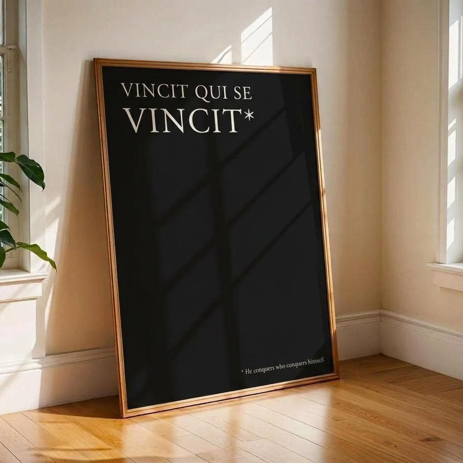A framed black poster leaning against a beige wall on a wooden floor in a sunlit room. The poster has white text reading 'VINCIT QUI SE VINCIT*' and is enclosed in a thin gold-toned frame.