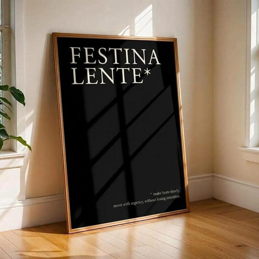 A framed black art print leaning against a beige wall on a wooden floor in a sunlit room. The print has the words 'FESTINA LENTE*' in large white serif lettering, with smaller white text near the bottom.
