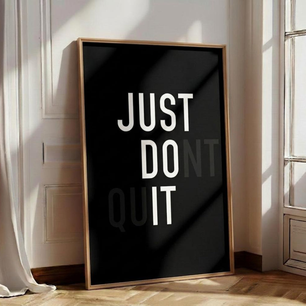 Framed poster with 'JUST DO IT' text on a black background in a room with light wood flooring and white curtains.