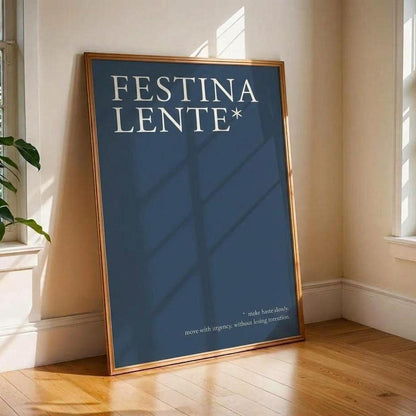 A framed art print with the text 'FESTINA LENTE*' in white on a dark blue background, leaning against a beige wall on a wooden floor in a sunlit room.