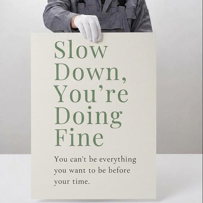 A person in a gray work jacket and white glove holds a large poster that reads 'Slow Down, You're Doing Fine' against a light gray background. The left side of the image features a beige panel with text describing professional giclée quality, including archival paper and pigment ink.