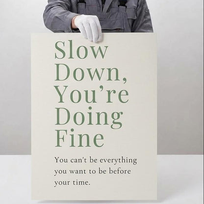 A person in a gray work jacket and white glove holds a large poster that reads 'Slow Down, You're Doing Fine' against a light gray background. The left side of the image features a beige panel with text describing professional giclée quality, including archival paper and pigment ink.