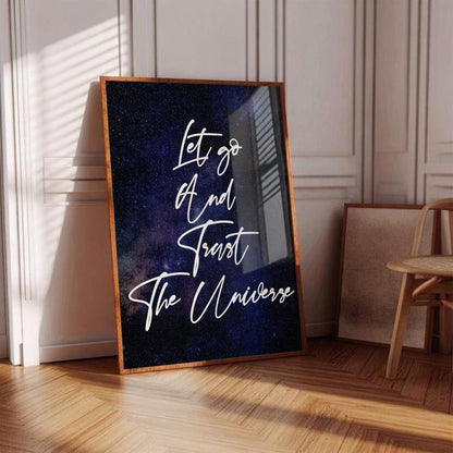 A framed art print leaning against a paneled wall on a wooden floor. The print features a dark blue and black galaxy background with white script text reading 'Let go And Trust The Universe'.