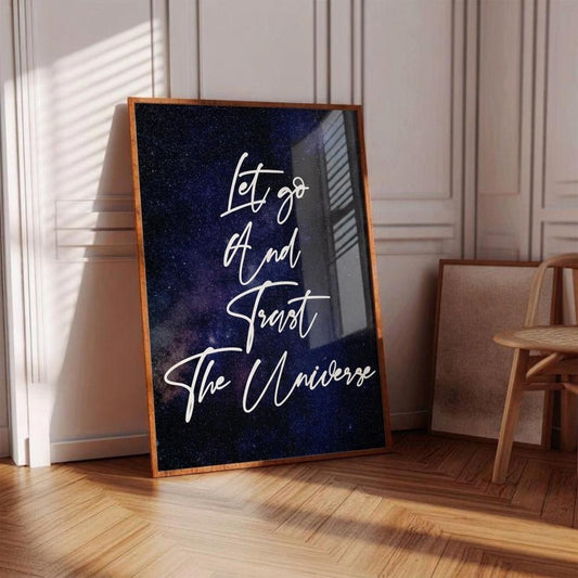 A framed art print leaning against a paneled wall on a wooden floor. The print features a dark blue and black galaxy background with white script text reading 'Let go And Trust The Universe'.