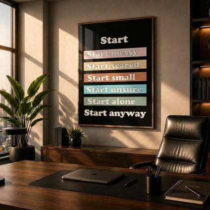 A framed motivational poster with the word 'Start' and stacked phrases like 'Start messy', 'Start scared', and 'Start anyway' in white text on colored bars, displayed on a wall in a modern office setting with a desk, chair, and city view.