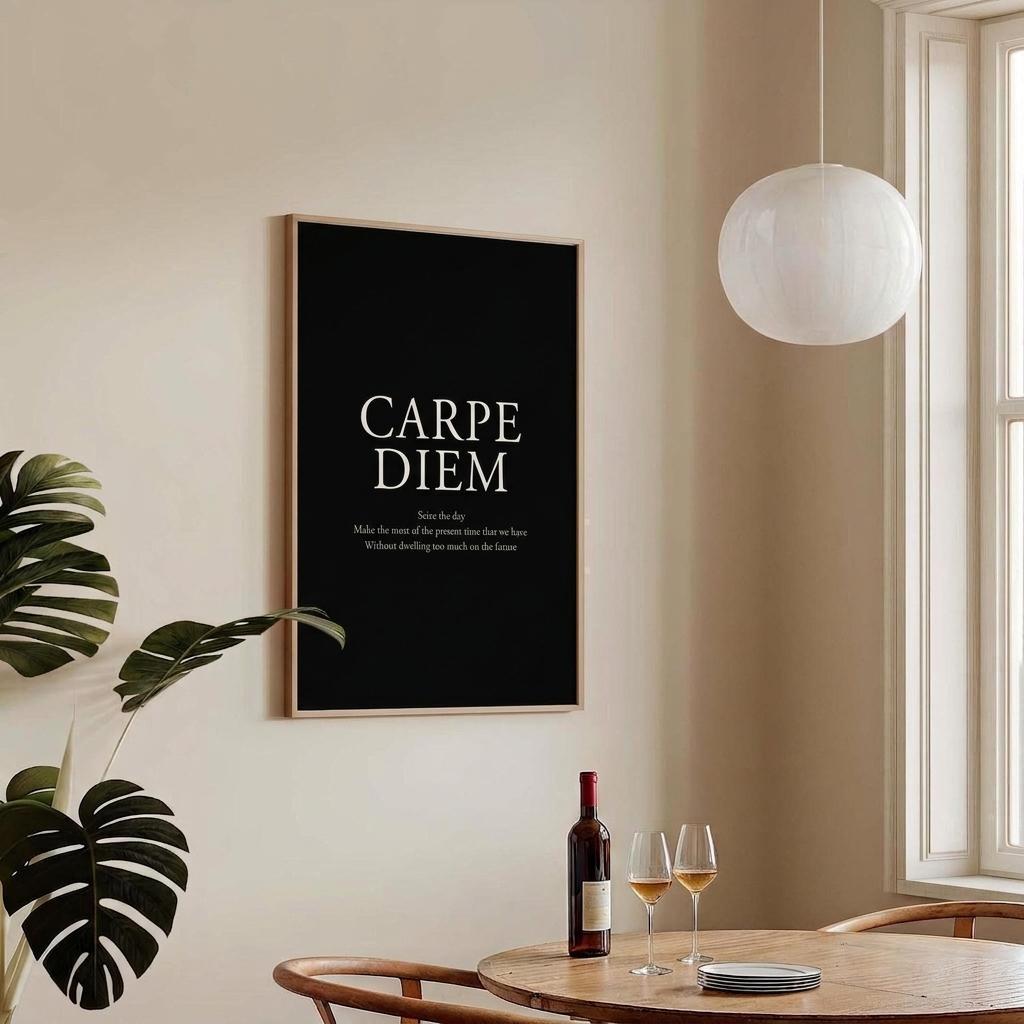 A framed black wall art print with the words 'CARPE DIEM' in white serif text hangs on a beige wall in a styled interior. A wooden dining table with wine glasses, a wine bottle, and chairs is below, with a white pendant light and window to the right.
