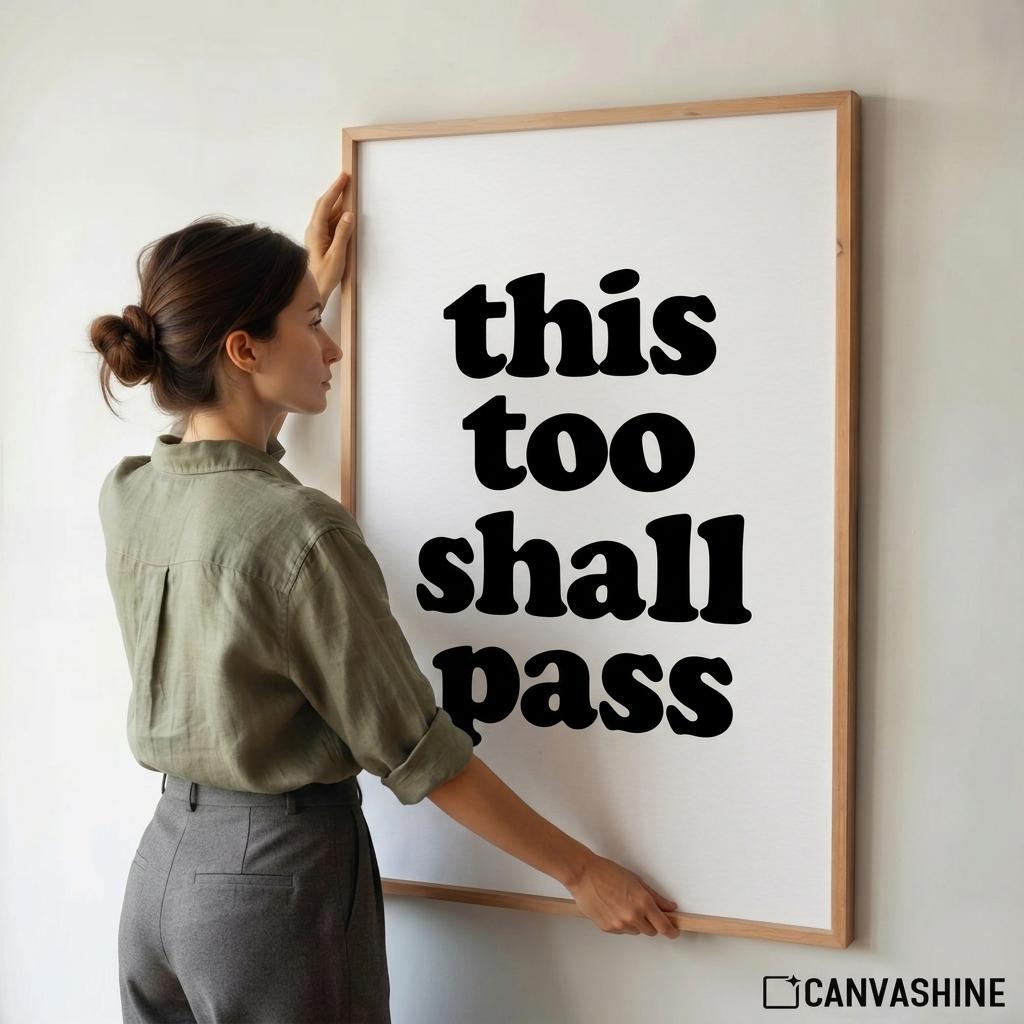 A woman in an olive green shirt and gray pants is adjusting a large framed wall art print that reads 'this too shall pass' in bold black text on a white background. The frame is light wood, and a CANVASHINE logo is visible in the bottom right corner.