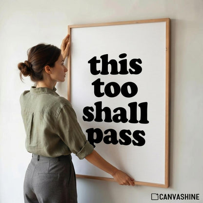 A woman in an olive green shirt and gray pants is adjusting a large framed wall art print that reads 'this too shall pass' in bold black text on a white background. The frame is light wood, and a CANVASHINE logo is visible in the bottom right corner.