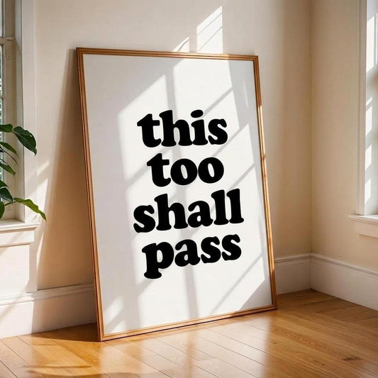 A framed typographic wall art print leaning against a beige wall on a wood floor, with sunlight streaming through nearby windows. The print has bold black text reading 'this too shall pass' on a white background with a thin warm-toned frame.