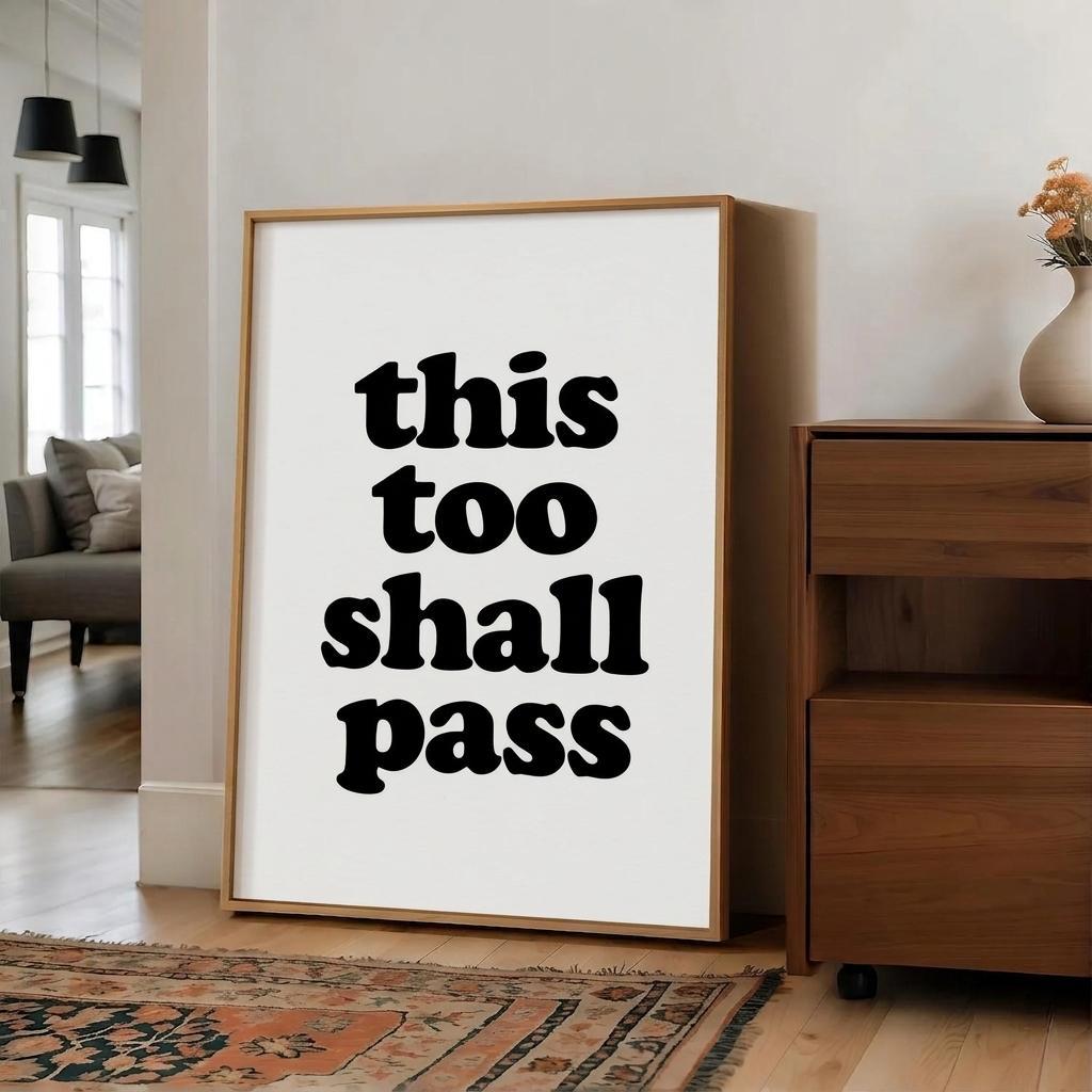 A framed typographic wall art print leaning against a white wall in a modern living room. The print has the phrase 'this too shall pass' in bold black lowercase letters on a white background, enclosed in a light natural wood frame.