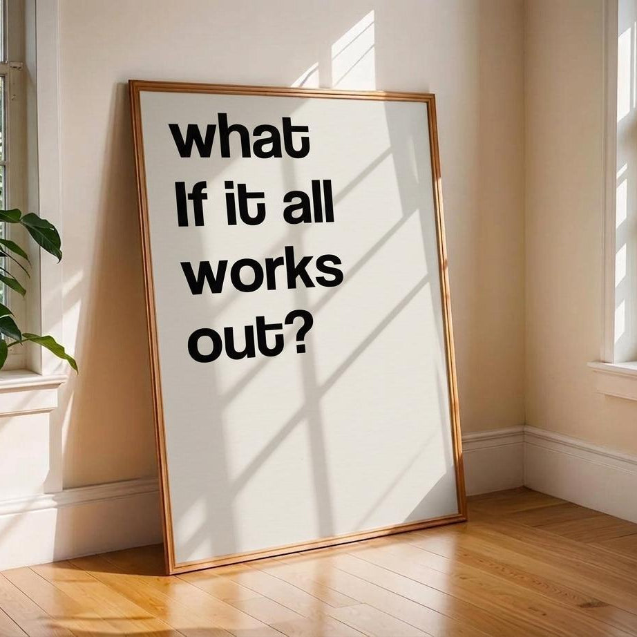A framed typographic wall art print leaning against a beige wall on a wood floor in a sunlit room. The print has large black text reading “what if it all works out?” on a white background with a thin warm-toned frame.