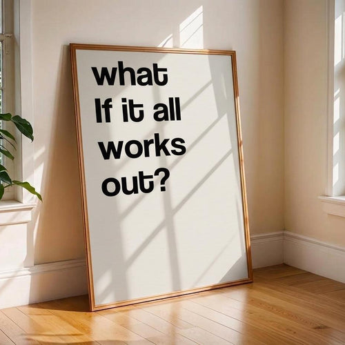 A framed typographic wall art print leaning against a beige wall on a wood floor in a sunlit room. The print has large black text reading “what if it all works out?” on a white background with a thin warm-toned frame.