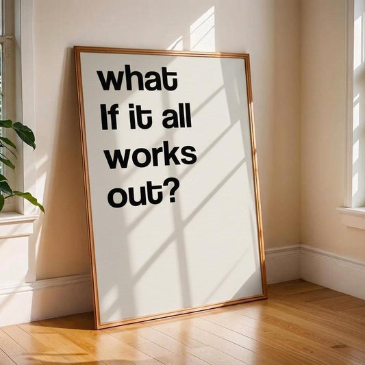 A framed typographic wall art print leaning against a beige wall on a wood floor in a sunlit room. The print has large black text reading “what if it all works out?” on a white background with a thin warm-toned frame.